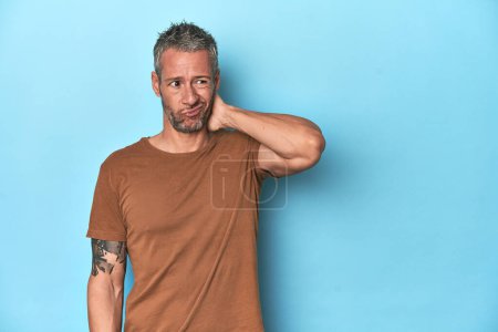 Photo for Middle-aged caucasian man on blue backdrop touching back of head, thinking and making a choice. - Royalty Free Image