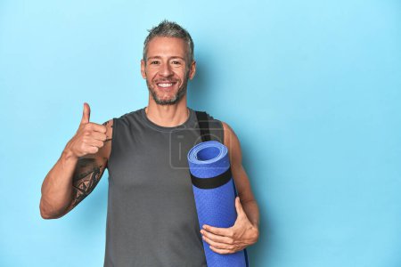 Photo for Athlete with a mat on a blue studio backdrop smiling and raising thumb up - Royalty Free Image