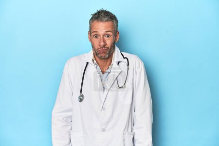 Photo for Caucasian middle-aged doctor on blue background shrugs shoulders and open eyes confused. - Royalty Free Image