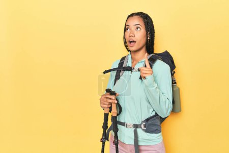 Téléchargez les photos : Femme indonésienne avec équipement de randonnée sur jaune pointant à l'envers avec bouche ouverte. - en image libre de droit