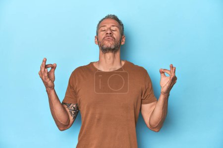 Photo for Middle-aged caucasian man on blue backdrop relaxes after hard working day, she is performing yoga. - Royalty Free Image
