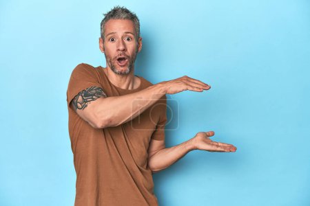 Photo for Middle-aged caucasian man on blue backdrop shocked and amazed holding a copy space between hands. - Royalty Free Image