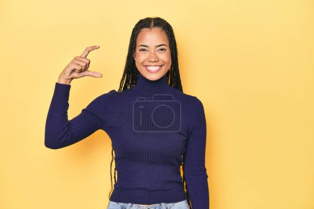 Photo for Young Indonesian woman on yellow studio backdrop holding something little with forefingers, smiling and confident. - Royalty Free Image