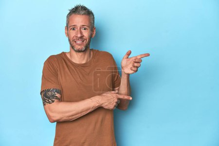 Foto de Hombre caucásico de mediana edad sobre fondo azul impactado señalando con los dedos índice a un espacio de copia. - Imagen libre de derechos