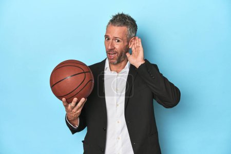 Photo for Middle-aged basketball coach on blue backdrop Middle-aged basketball coach on blue backdroptrying to listening a gossip. - Royalty Free Image
