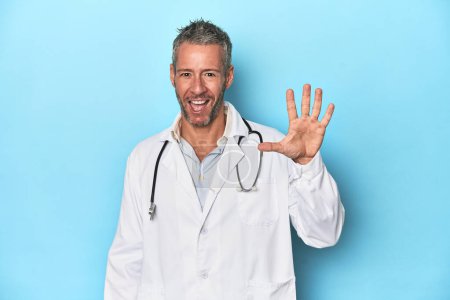 Foto de Caucásico médico de mediana edad sobre fondo azul sonriente alegre mostrando número cinco con los dedos. - Imagen libre de derechos