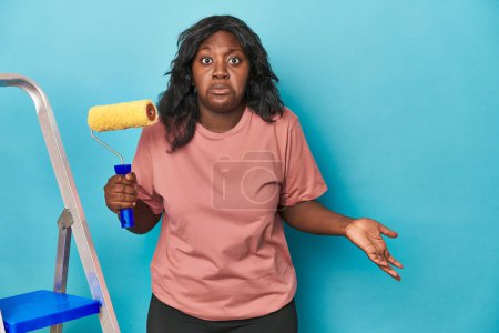 Photo for Curvy woman with paint roller and ladder shrugs shoulders and open eyes confused. - Royalty Free Image