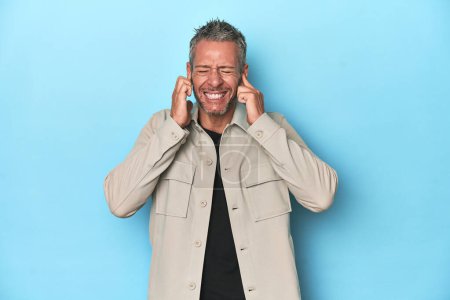 Photo for Middle-aged caucasian man on blue backdrop covering ears with hands. - Royalty Free Image