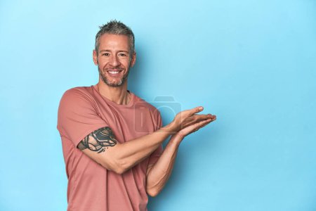 Photo for Middle-aged caucasian man on blue backdrop holding a copy space on a palm. - Royalty Free Image