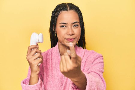 Foto de Mujer indonesia con limpiador facial en amarillo apuntando con el dedo a usted como si invitando a acercarse. - Imagen libre de derechos