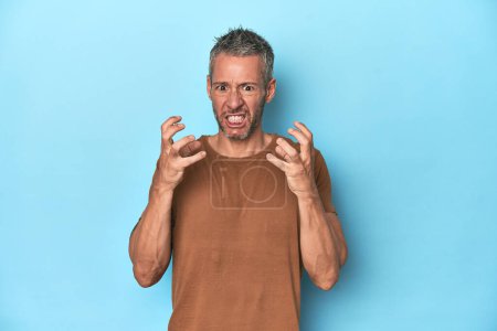 Foto de Hombre caucásico de mediana edad sobre fondo azul molesto gritando con las manos tensas. - Imagen libre de derechos