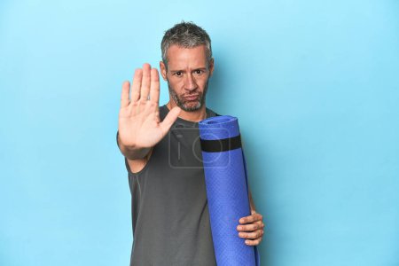 Photo for Athlete with a mat on a blue studio backdrop standing with outstretched hand showing stop sign, preventing you. - Royalty Free Image