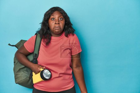 Photo for Curvy explorer with backpack and flashlight shrugs shoulders and open eyes confused. - Royalty Free Image