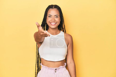 Photo for Young Indonesian woman on yellow studio backdrop stretching hand at camera in greeting gesture. - Royalty Free Image