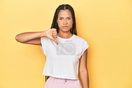 Téléchargez les photos : Jeune femme indonésienne sur fond de studio jaune montrant pouce baissé, concept de déception. - en image libre de droit