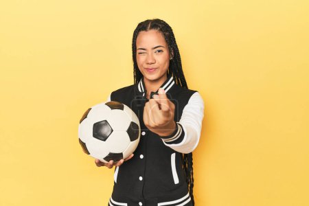 Photo for Indonesian schoolgirl with soccer ball on yellow pointing with finger at you as if inviting come closer. - Royalty Free Image