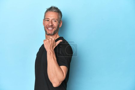 Photo for Middle-aged caucasian man on blue backdrop points with thumb finger away, laughing and carefree. - Royalty Free Image