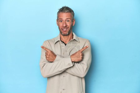 Photo for Middle-aged caucasian man on blue backdrop points sideways, is trying to choose between two options. - Royalty Free Image