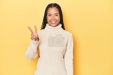 Photo for Young Indonesian woman on yellow studio backdrop showing victory sign and smiling broadly. - Royalty Free Image