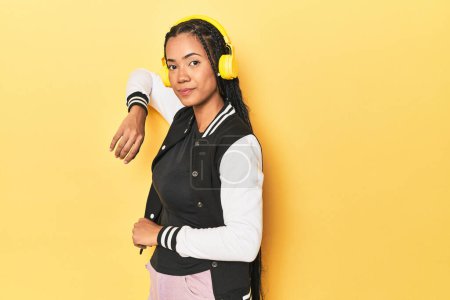Téléchargez les photos : Femme indonésienne en veste de baseball avec écouteurs sur fond de studio jaune - en image libre de droit