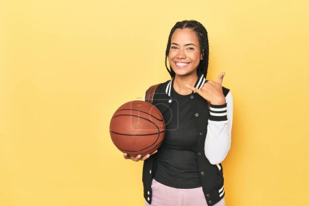 Photo for Indonesian schoolgirl with basketball on yellow showing a mobile phone call gesture with fingers. - Royalty Free Image