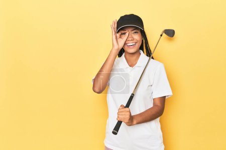 Photo for Indonesian female golfer on yellow backdrop excited keeping ok gesture on eye. - Royalty Free Image