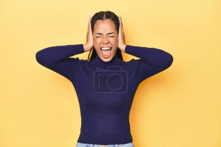 Photo for Young Indonesian woman on yellow studio backdrop covering ears with hands trying not to hear too loud sound. - Royalty Free Image