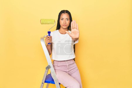 Téléchargez les photos : Femme indonésienne avec rouleau sur l'échelle sur jaune debout avec la main tendue montrant le signe d'arrêt, vous empêchant. - en image libre de droit