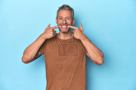 Téléchargez les photos : Homme caucasien d'âge moyen sur fond bleu sourit, les doigts pointant vers la bouche. - en image libre de droit