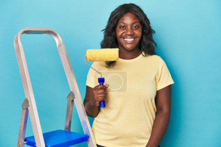 Photo for Curvy woman with ladder and paint roller on a blue backdrop - Royalty Free Image