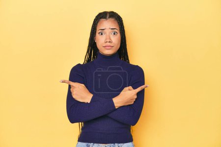 Téléchargez les photos : Jeune femme indonésienne sur fond de studio jaune points de côté, essaie de choisir entre deux options. - en image libre de droit