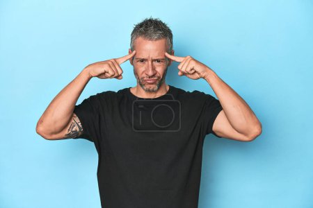 Photo for Middle-aged caucasian man on blue backdrop focused on a task, keeping forefingers pointing head. - Royalty Free Image
