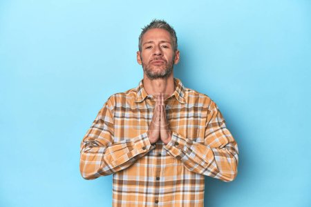 Foto de Hombre caucásico de mediana edad sobre fondo azul rezando, mostrando devoción, persona religiosa buscando inspiración divina. - Imagen libre de derechos