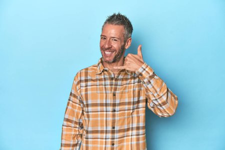 Photo for Middle-aged caucasian man on blue backdrop showing a mobile phone call gesture with fingers. - Royalty Free Image