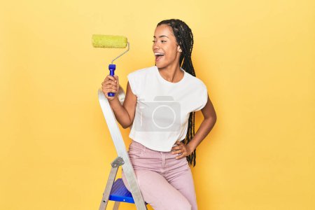 Photo for Indonesian woman with roller on ladder on yellow points with thumb finger away, laughing and carefree. - Royalty Free Image