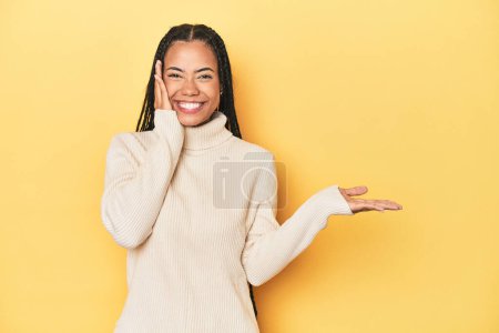 Photo for Young Indonesian woman on yellow studio backdrop holds copy space on a palm, keep hand over cheek. Amazed and delighted. - Royalty Free Image
