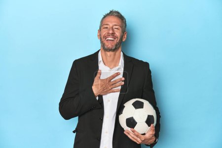 Foto de Entrenador de fútbol caucásico, de mediana edad en el conjunto azul se ríe en voz alta manteniendo la mano en el pecho. - Imagen libre de derechos