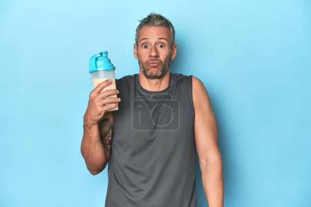 Photo for Athlete holding protein shake on blue backdrop shrugs shoulders and open eyes confused. - Royalty Free Image