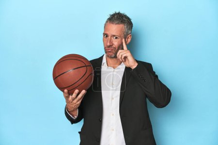 Photo for Middle-aged basketball coach on blue backdrop Middle-aged basketball coach on blue backdroppointing temple with finger, thinking, focused on a task. - Royalty Free Image