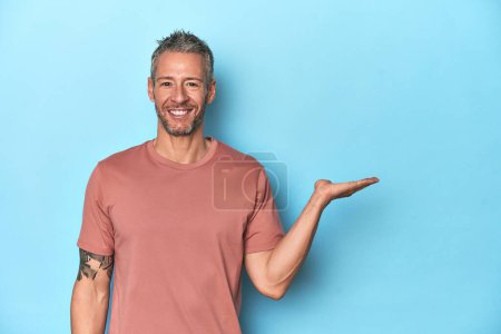 Photo for Middle-aged caucasian man on blue backdrop showing a copy space on a palm and holding another hand on waist. - Royalty Free Image