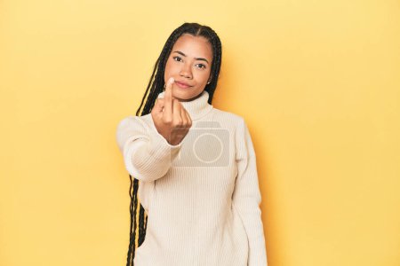 Photo for Young Indonesian woman on yellow studio backdrop pointing with finger at you as if inviting come closer. - Royalty Free Image