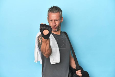 Photo for Athlete with gym backpack on blue background showing fist to camera, aggressive facial expression. - Royalty Free Image