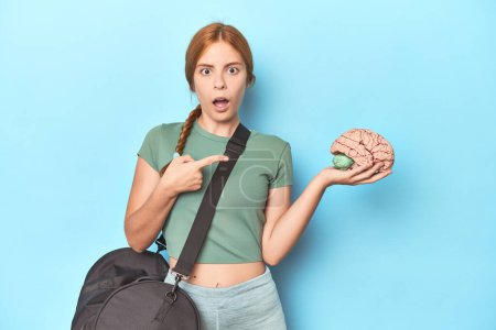 Photo for Sporty redhead with brain model on blue background pointing to the side - Royalty Free Image