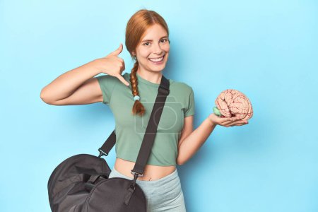 Photo for Sporty redhead with brain model on blue background showing a mobile phone call gesture with fingers. - Royalty Free Image