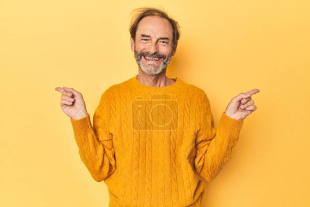 Photo for Caucasian middle-aged man in yellow studio pointing to different copy spaces, choosing one of them, showing with finger. - Royalty Free Image