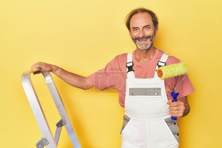 Téléchargez les photos : Homme d'âge moyen peint un mur jaune avec échelle - en image libre de droit
