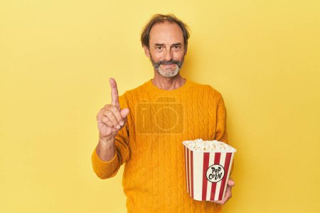 Téléchargez les photos : Homme appréciant pop-corn en studio jaune montrant numéro un avec doigt. - en image libre de droit