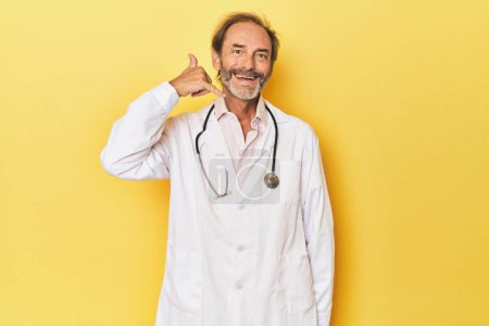 Téléchargez les photos : Médecin avec stéthoscope en studio jaune montrant un geste d'appel téléphonique mobile avec les doigts. - en image libre de droit