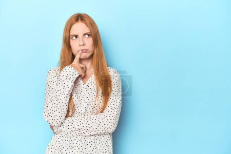 Redhead young woman on blue background looking sideways with doubtful and skeptical expression.