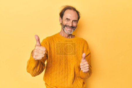 Foto de Caucásico hombre de mediana edad en estudio amarillo levantando ambos pulgares hacia arriba, sonriendo y confiado. - Imagen libre de derechos